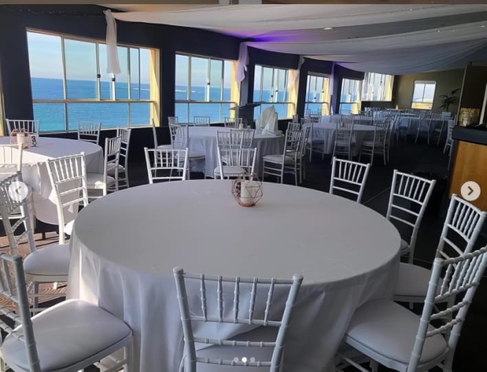 A room with white tables and chairs and a view of the ocean.