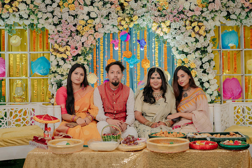 Engagement party with a couple and guests seated in front of a vibrant flower backdrop, surrounded by colorful decorations and traditional festive food.