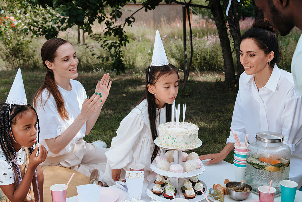 Outdoor birthday celebration with a young girl blowing out candles on a cake, surrounded by friends and family wearing party hats, with cupcakes and drinks on the table.