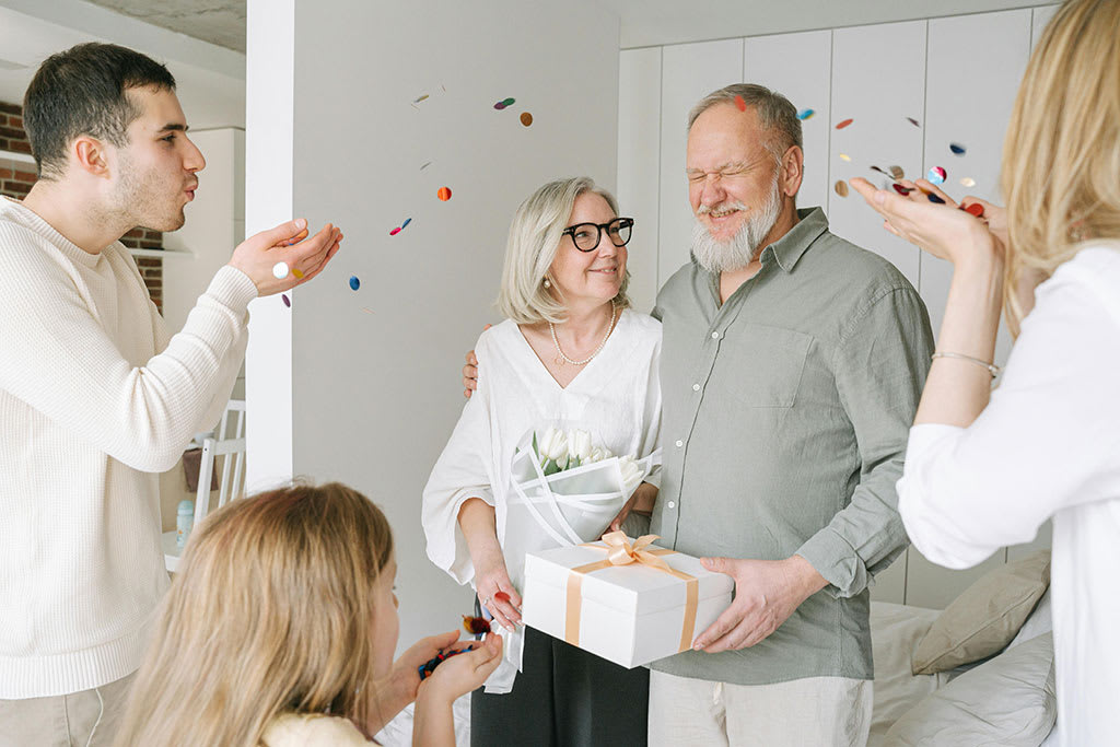 Family celebrating an anniversary at home with confetti, an elderly couple holding flowers and a wrapped gift, surrounded by smiling relatives in a warm and joyful atmosphere.