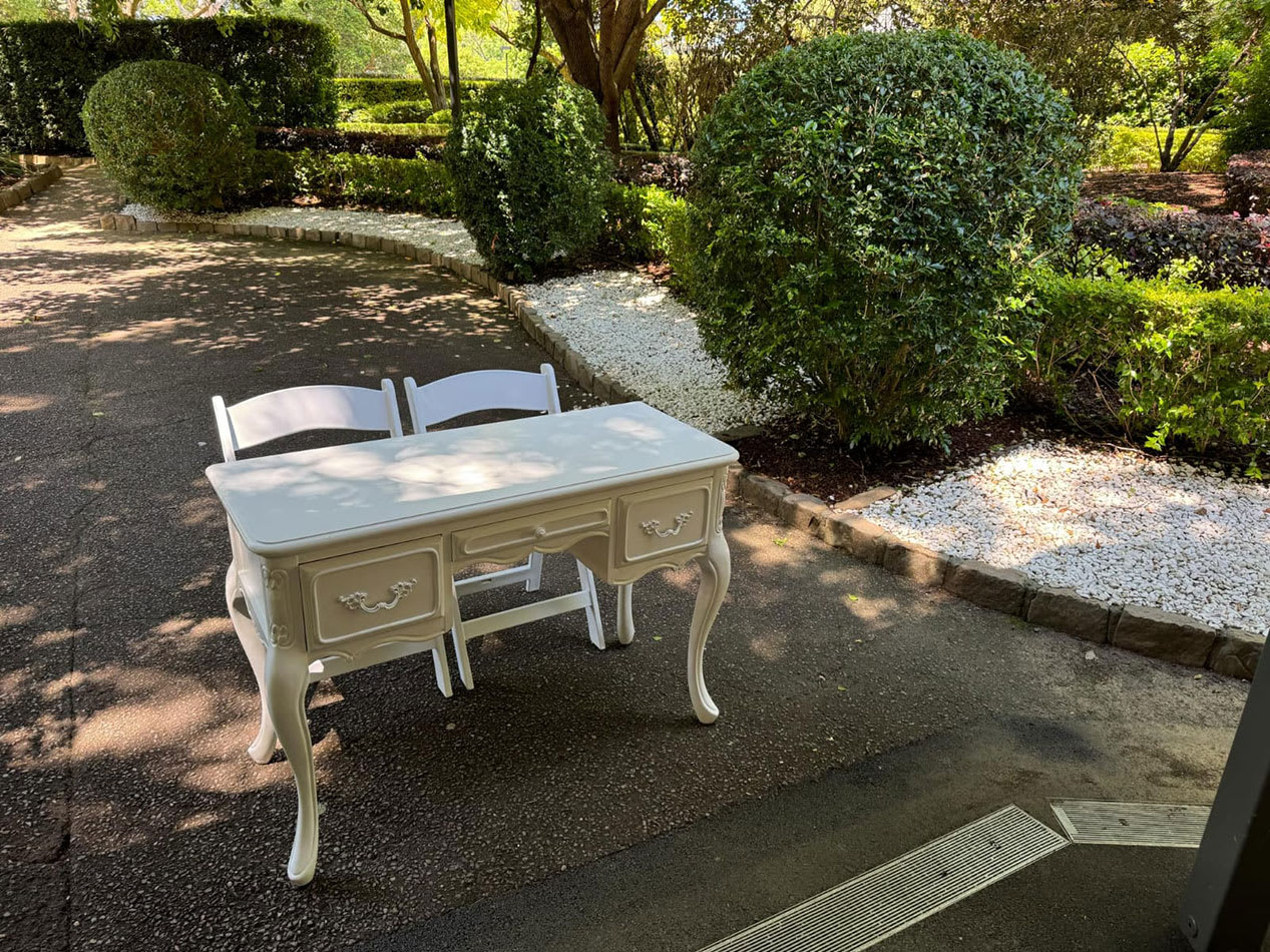 Ornate vintage style table with classic drawer detailing, placed with two folding white chairs on a shaded garden pathway lined with shrubs and white gravel.