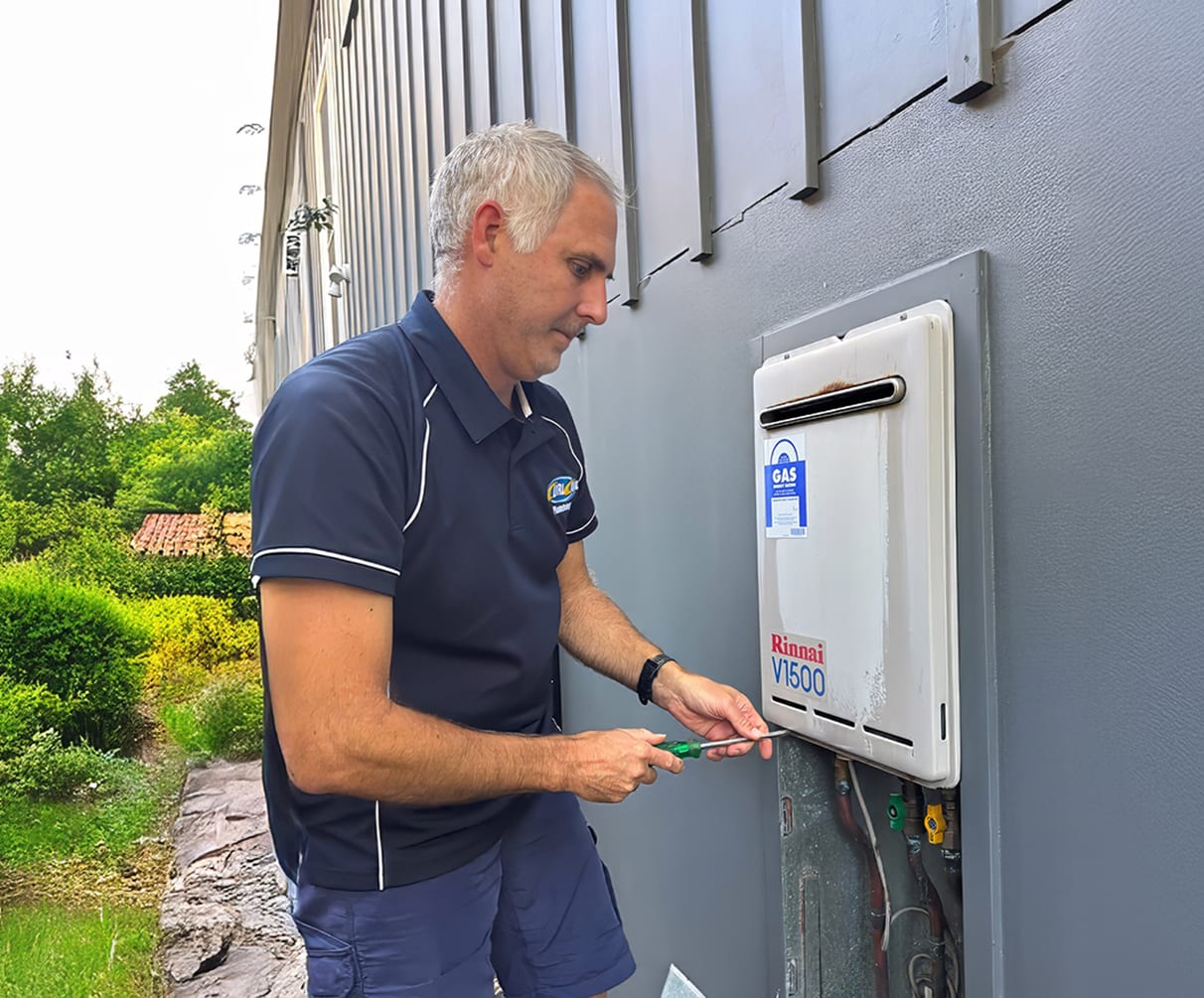 Licensed plumber servicing an outdoor wall-mounted gas hot water system on the side of a residential building with garden landscaping nearby
