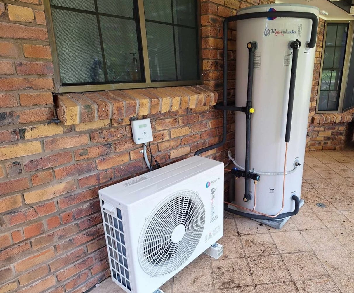 Outdoor heat pump hot water installation with a white compressor unit, vertical storage tank, insulated pipework, and brick exterior wall on a tiled patio