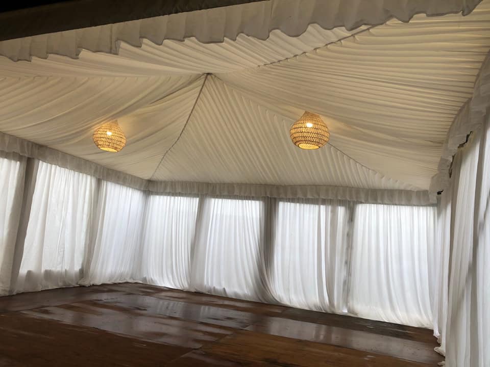 A white tent with white curtains and a wooden floor illuminated by a white pendant rope light.