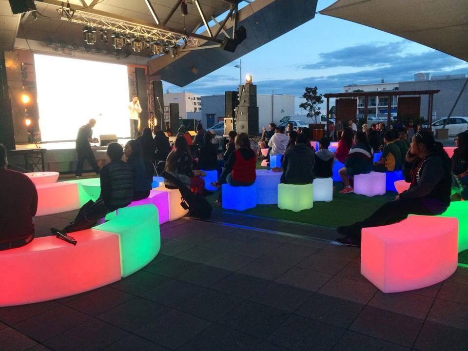 A group of people sitting on a bench in front of a Glow Cube Hire screen.