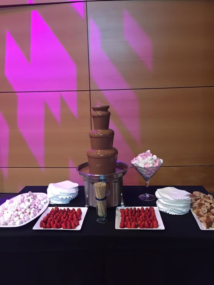 A chocolate fountain on a table with 1kg of strawberries and desserts on it.