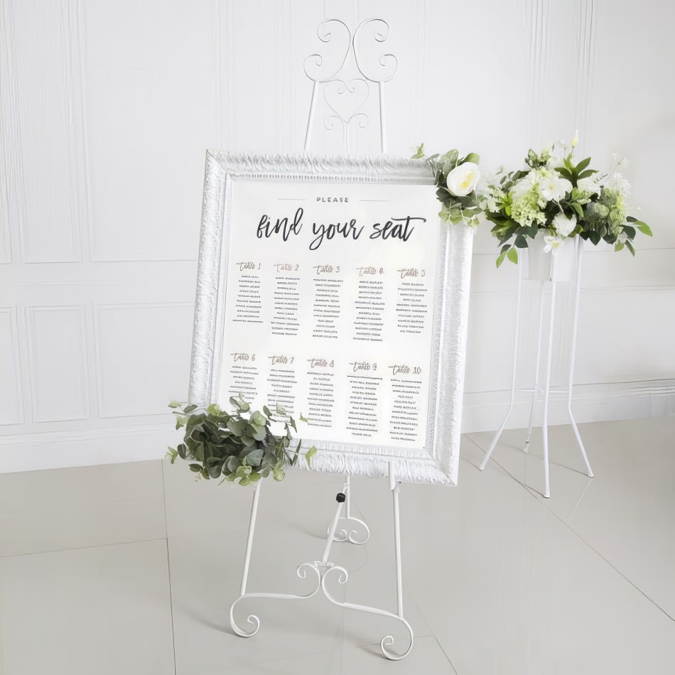 A wedding seating chart on a white French easel in front of flowers.