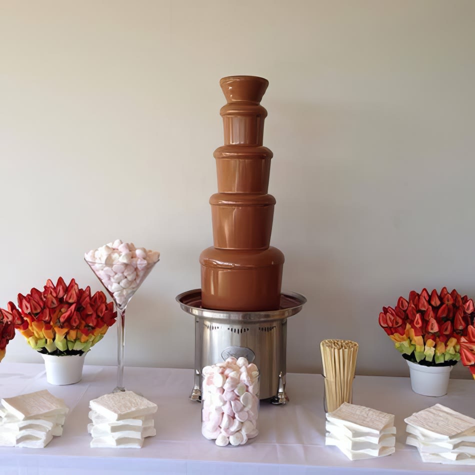 A large commercial chocolate fountain with marshmallows and flowers on a table.