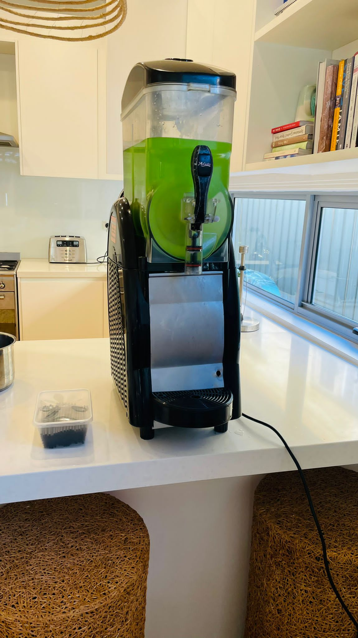 A green juice machine sitting on a counter in a kitchen, available for Slushie Machine Hire: Pkg 1, capable of making up to *60 drinks*.