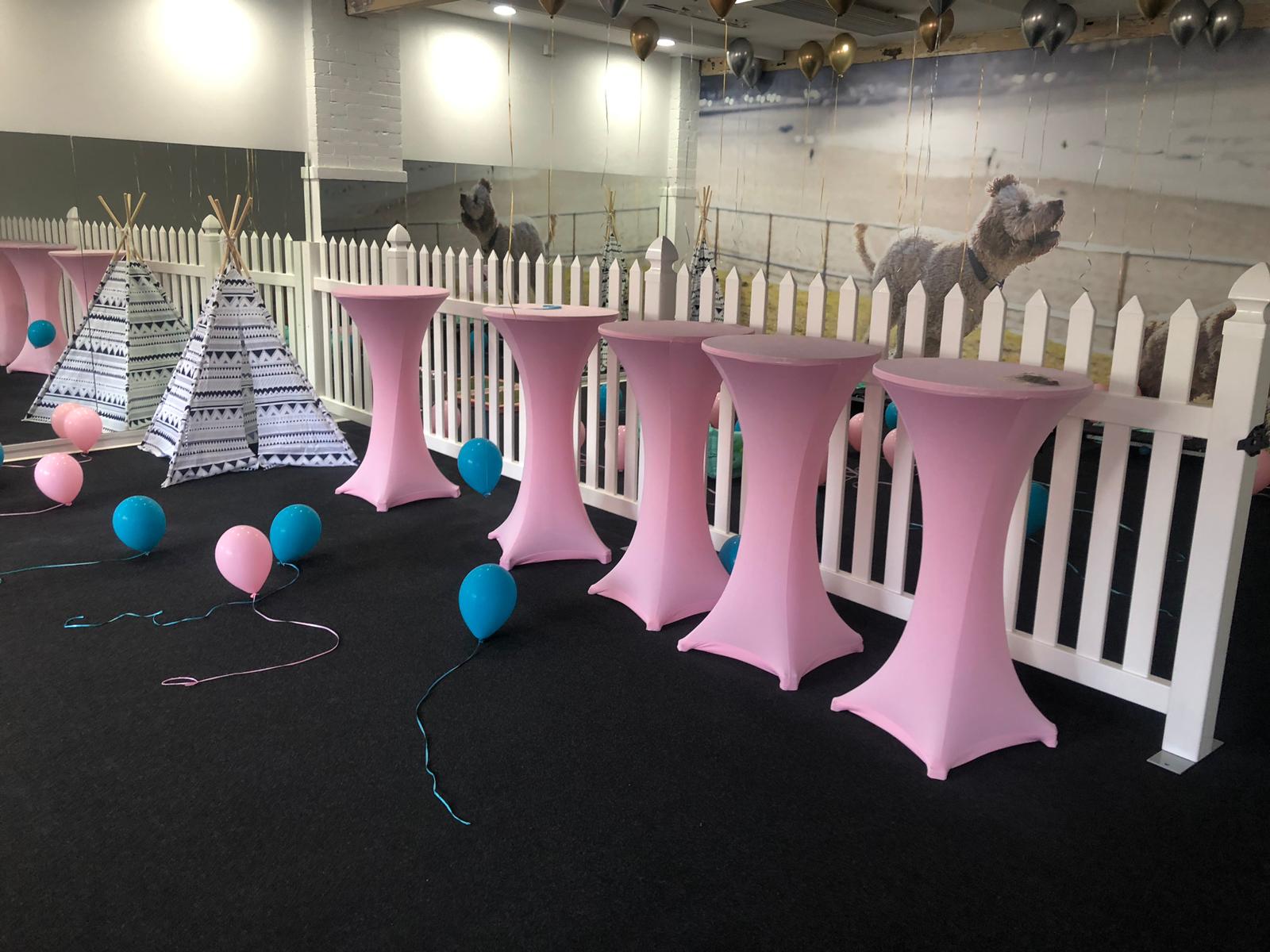 Pink balloons and tables in a room with a white picket fence, equipped with a stainless steel cocktail bar table.