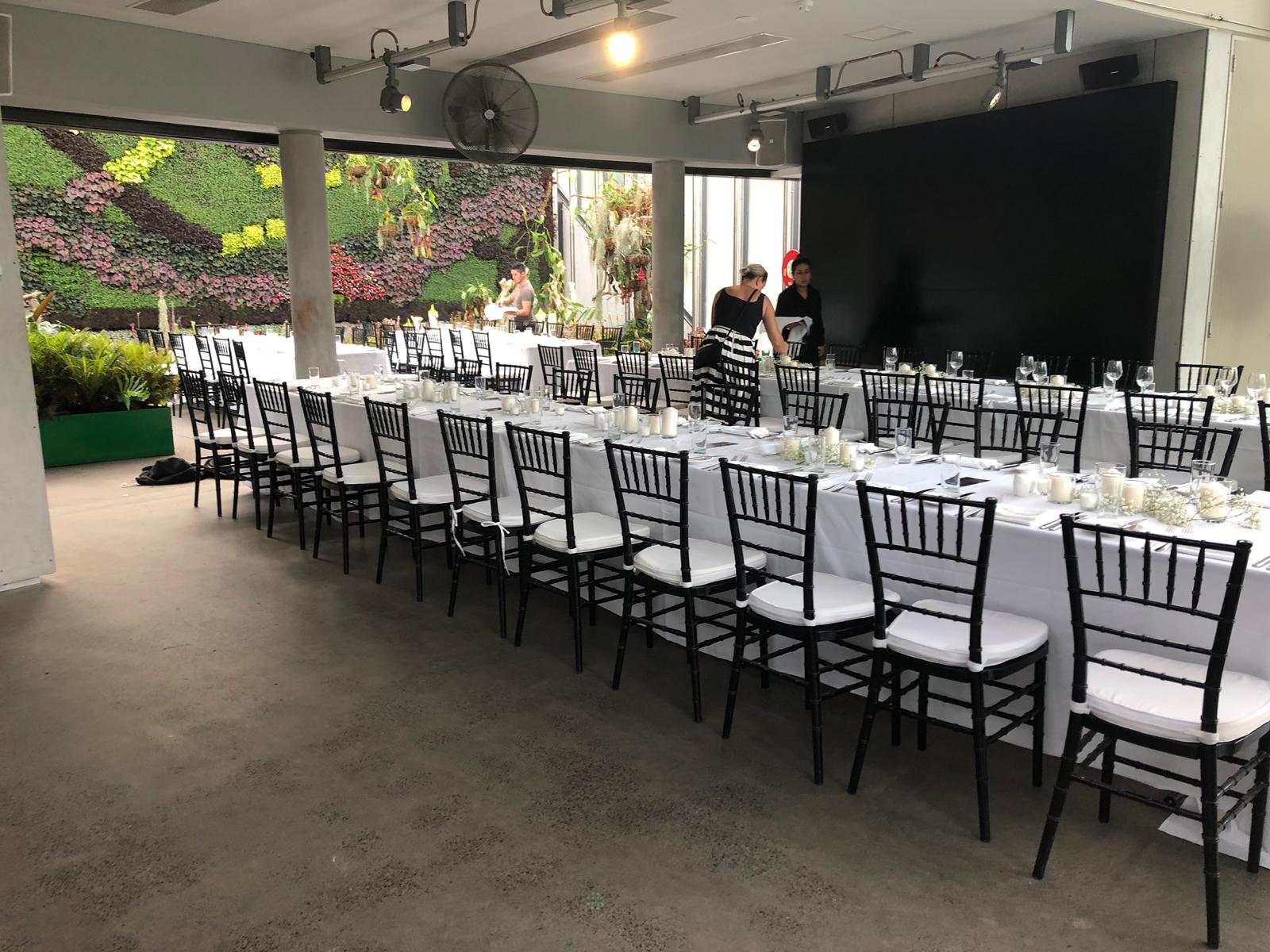 A long table set up with black Tiffany chairs and white tablecloths.