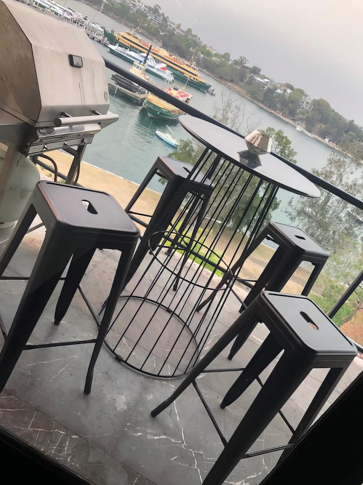 A black wire cocktail table with stools and a grill on a balcony overlooking the water.