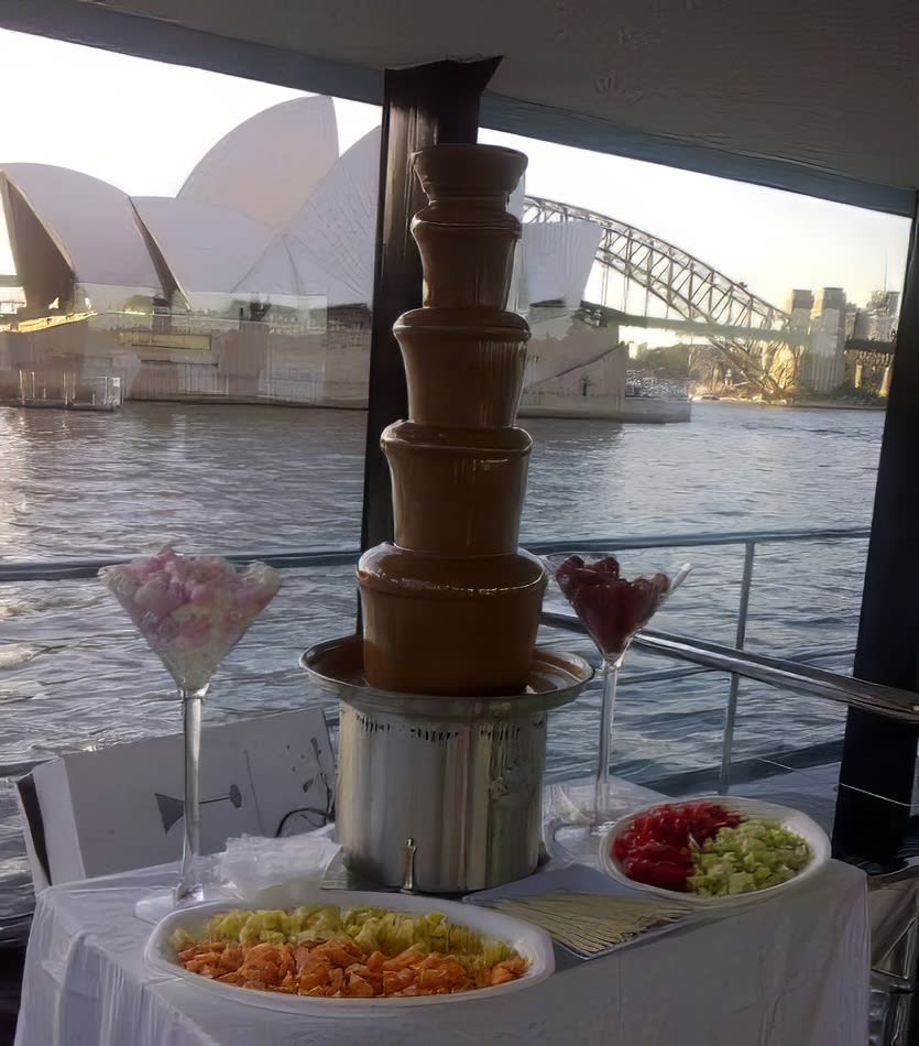 A chocolate fountain on a boat with a large fruit platter and a view of the Sydney Opera House, showcasing in season fruits.