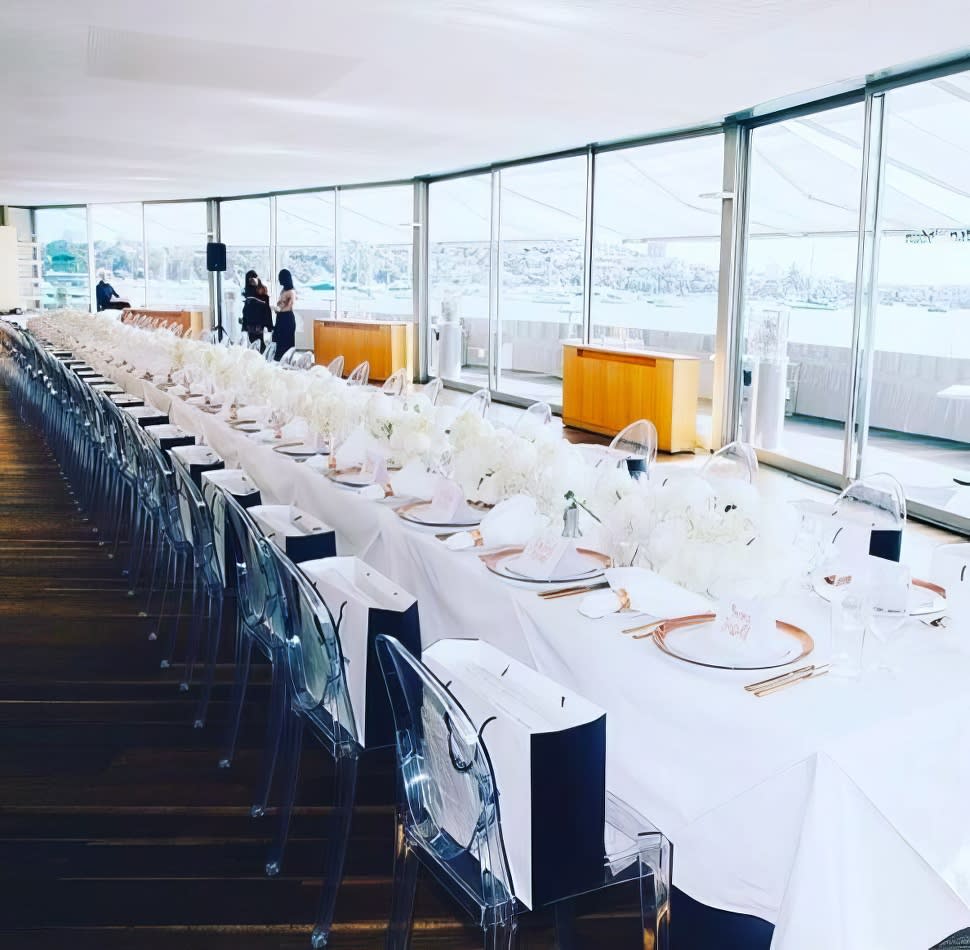 A long white table with white chairs and a view of the harbour.