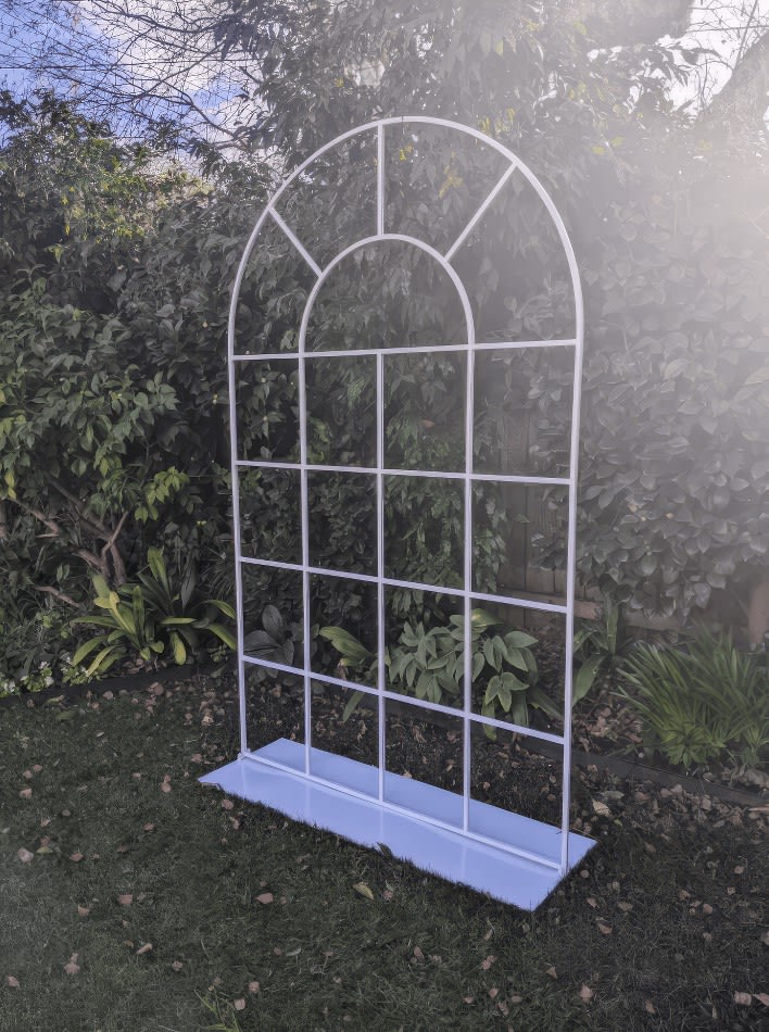 A white arbour, resembling a French window arch frame, situated in the middle of a garden.