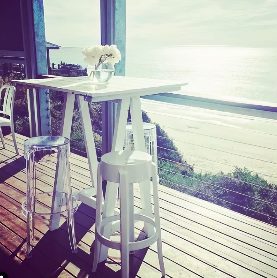 A white table and ivory stools on a deck overlooking the ocean.