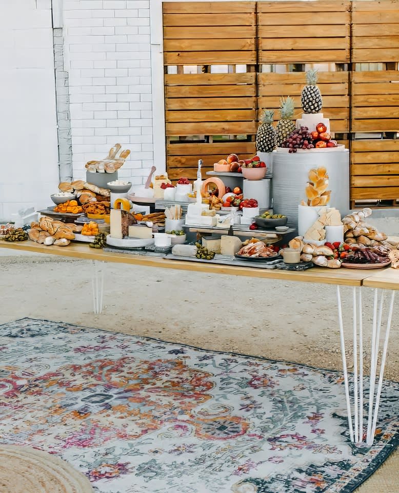 A White Hairpin Banquet Table with a Timber Top covered in a variety of delicious food, set on a rug in front of a brick wall.