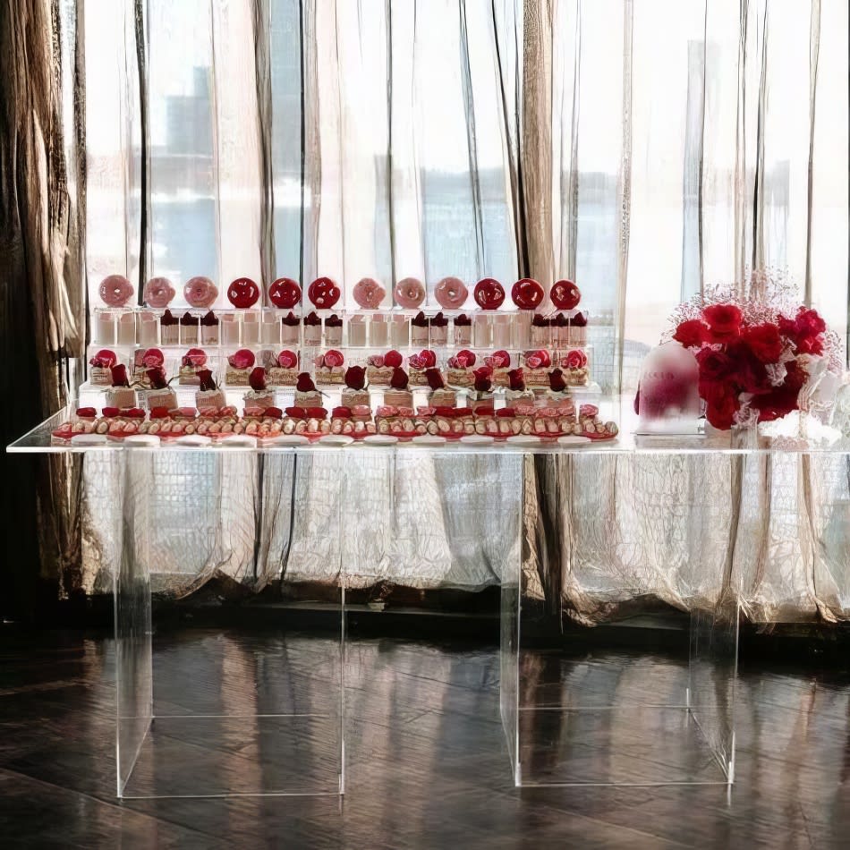 A clear acrylic table with cupcakes and red roses in front of a window.