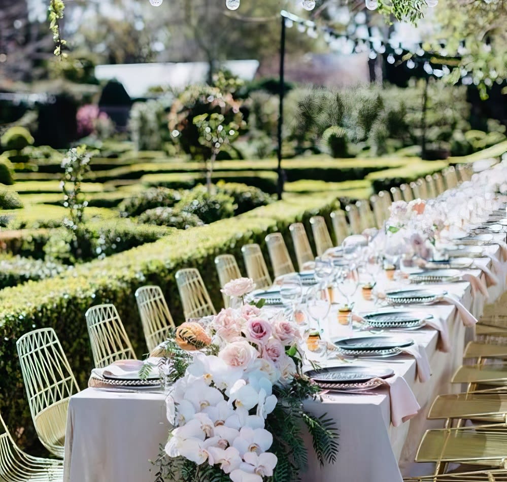 A long table set up in a garden with gold wire chairs.