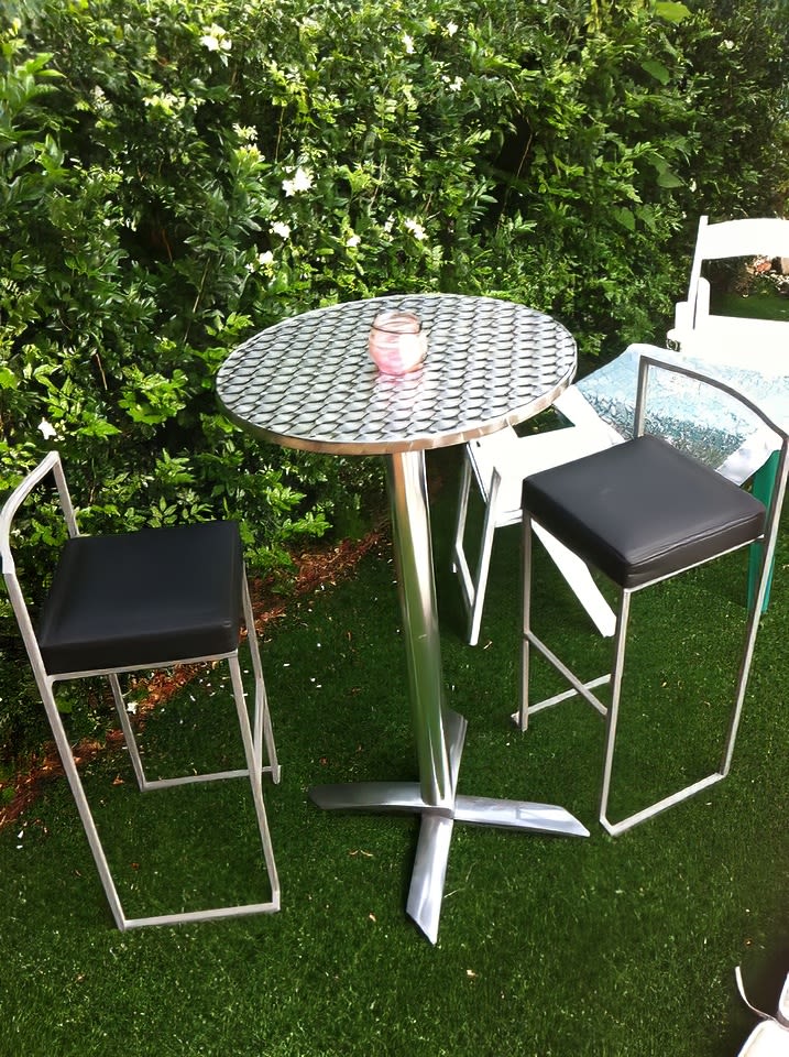 A stainless steel cocktail bar table and two stools in a yard.