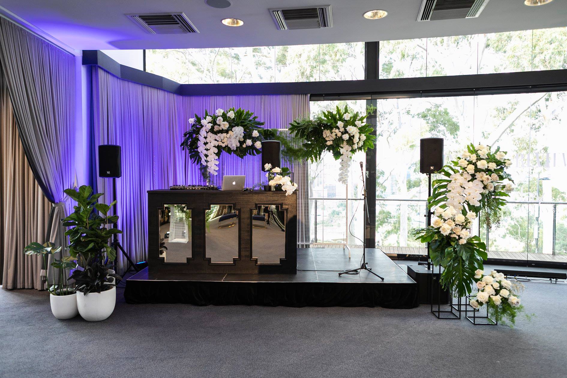 A room adorned with flowers and plants featuring a PA system with wireless mic and speaker stands.