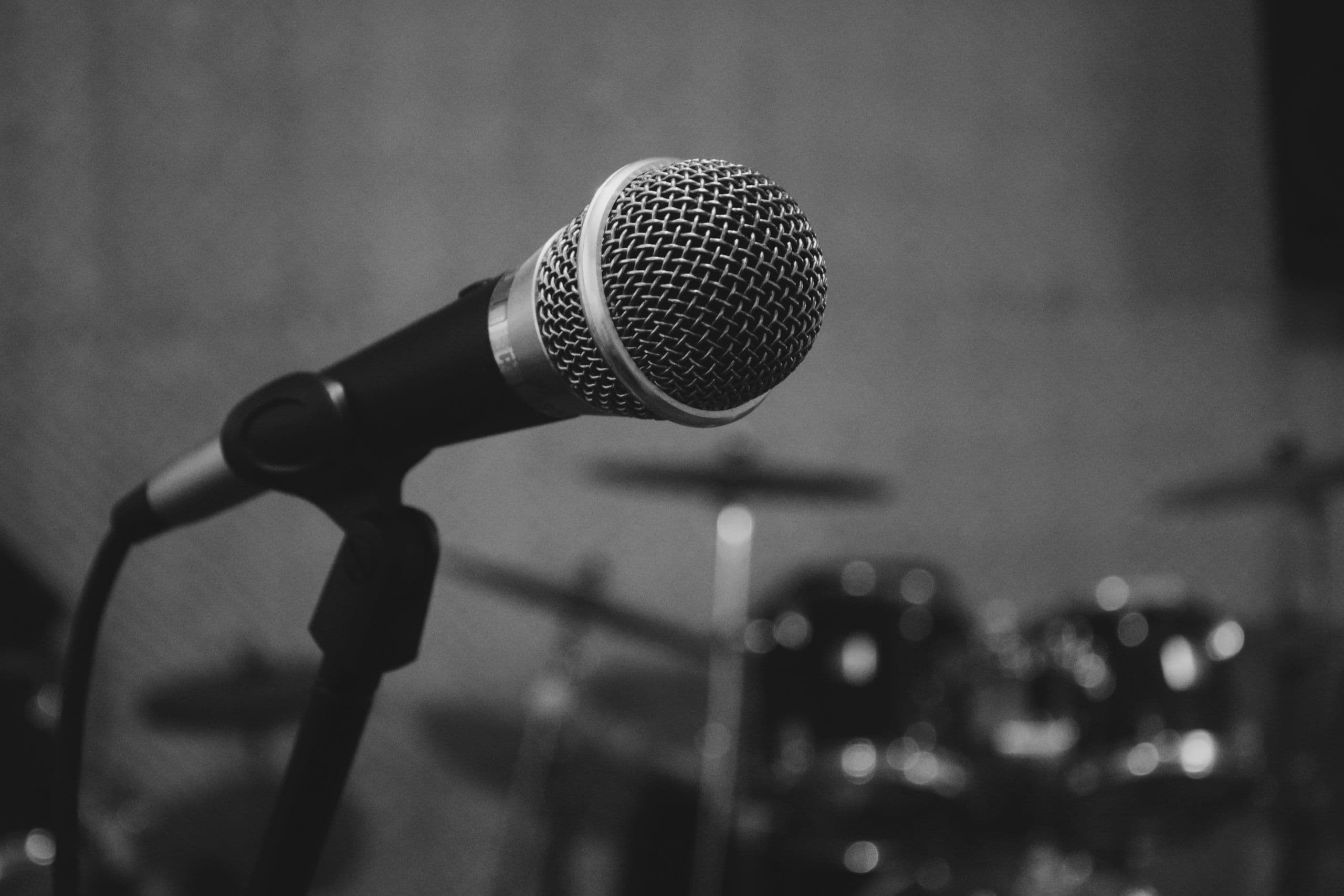 A black and white photo of a corded microphone in front of a drum set.