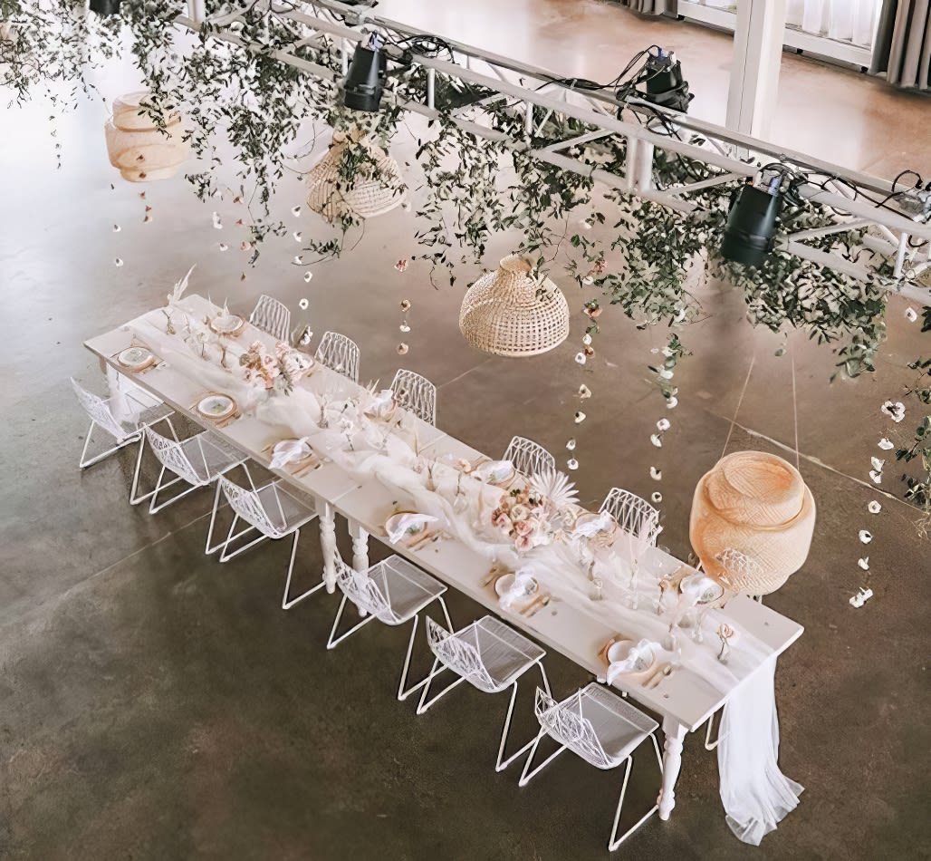 A white table with white chairs adorned with greenery hanging from the ceiling.