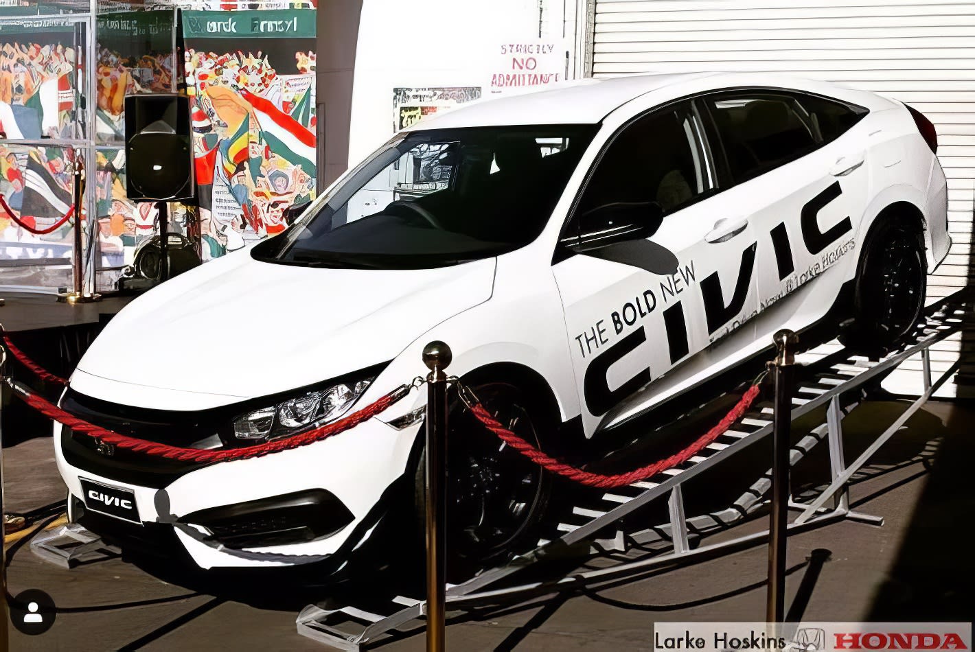 A white Honda Civic is on display in front of a crowd of people, surrounded by gold bollards.