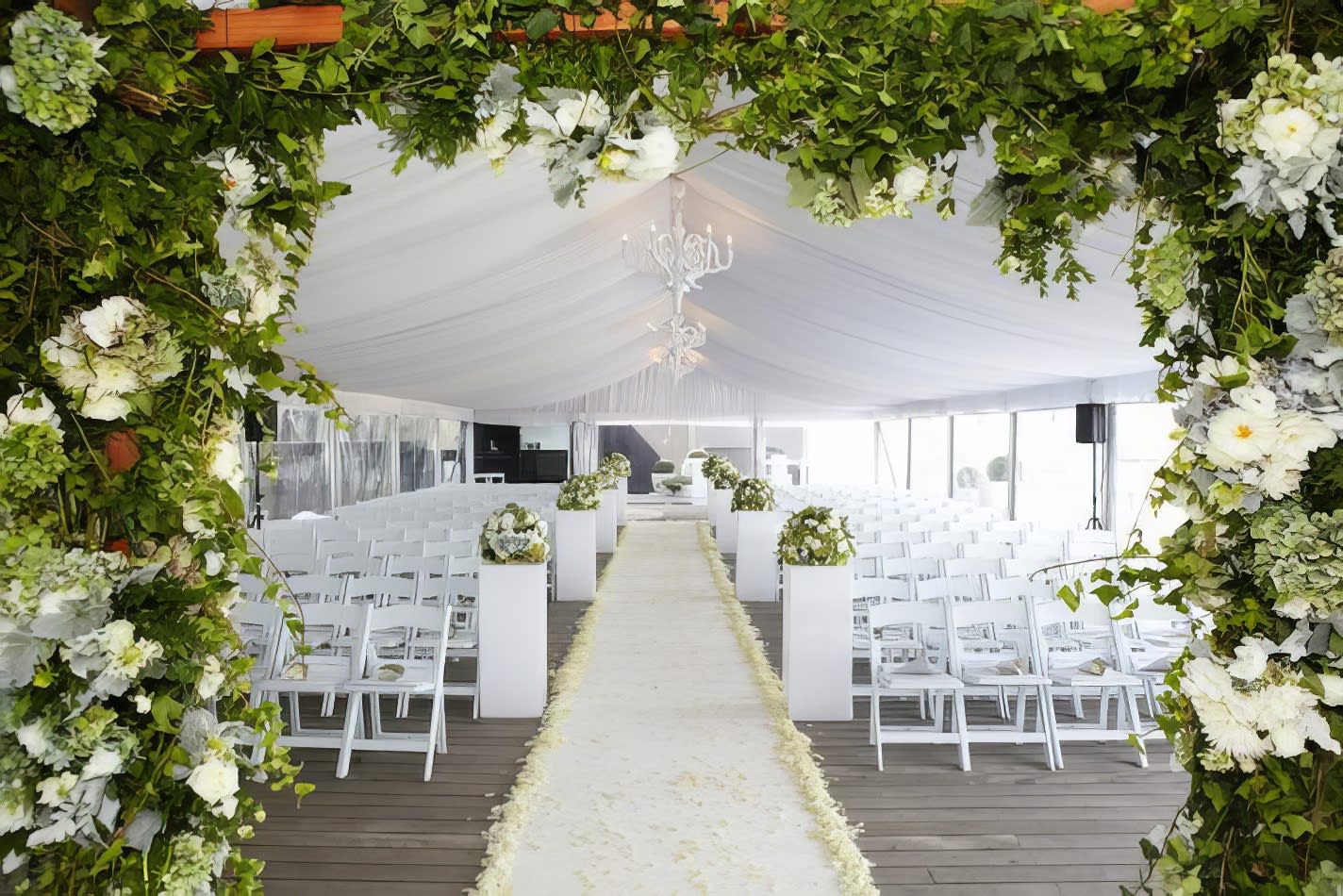 A wedding ceremony set up in a tent with white flowers and greenery, featuring elegant White Padded Folding Chairs (Gladiator chair) for seating.