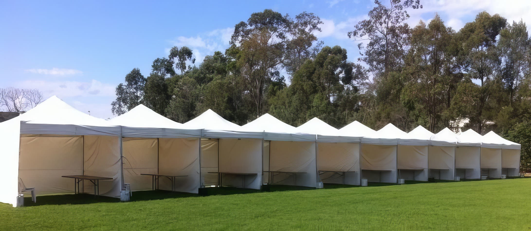 A row of white 3mx3m pop up marquees with walls on 3 sides, set up on a grassy field.