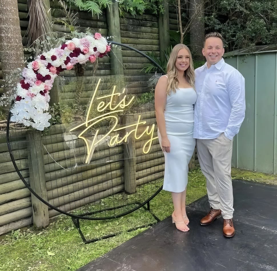 A man and woman posing joyfully next to a neon "Let's Party" sign.