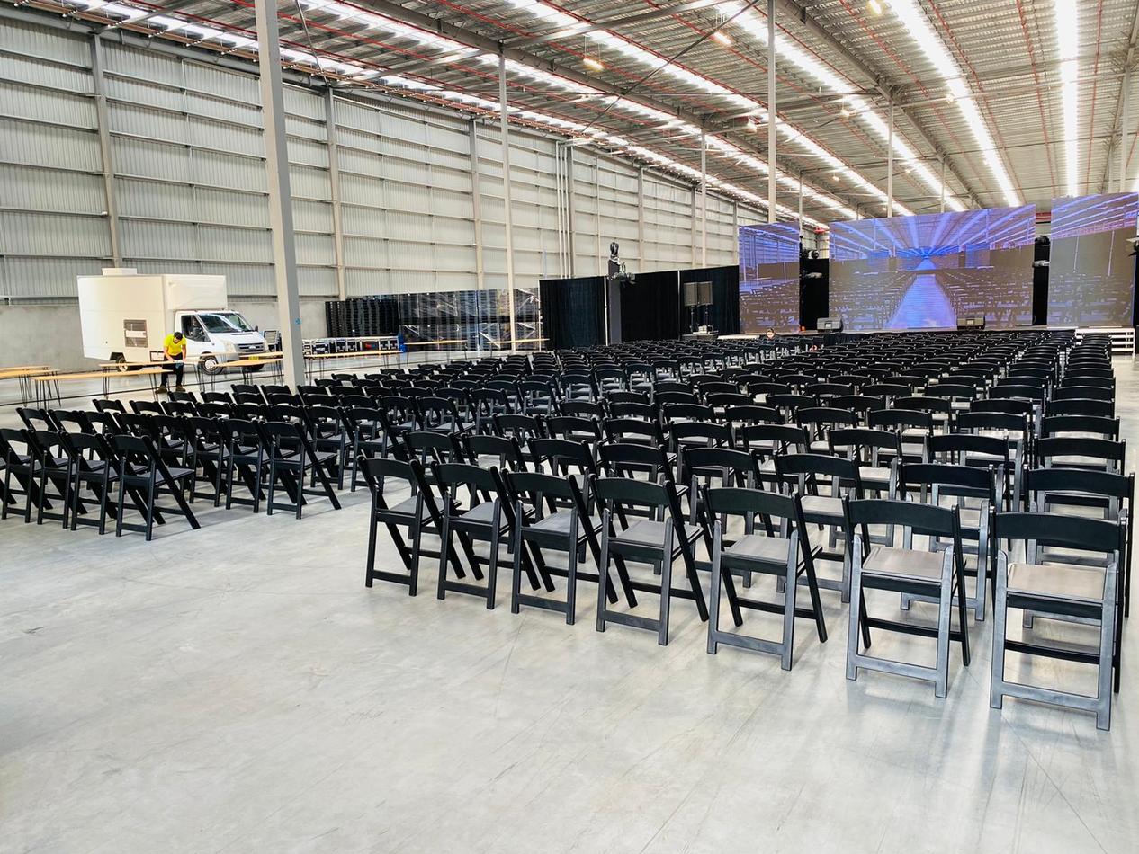 A large warehouse with rows of black padded folding chairs and a large screen.