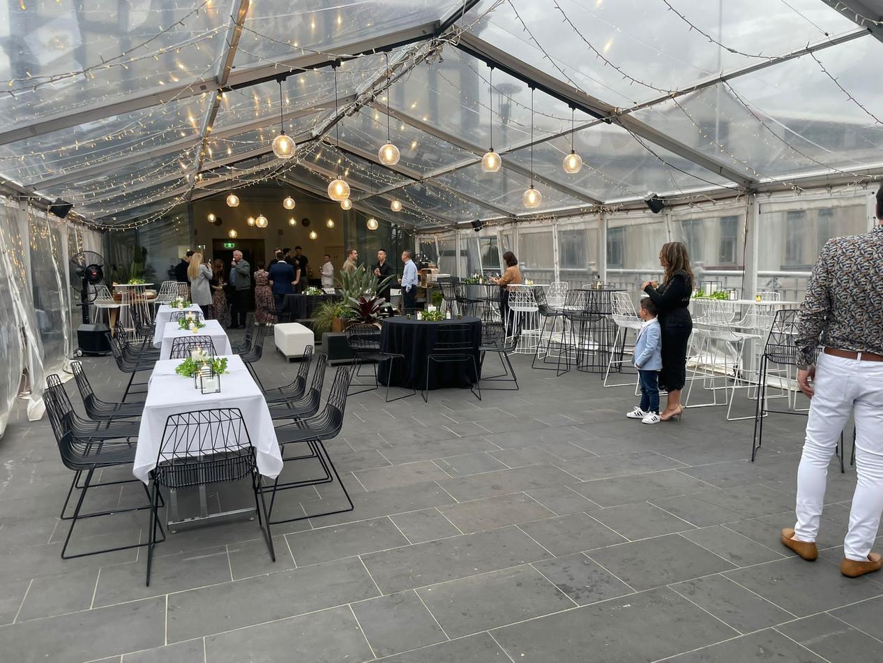 A clear tent with tables and black wire chairs in it.