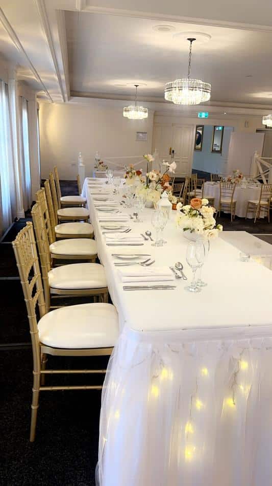 A table set up for a wedding with white linens and Gold Tiffany Chair Hire, adorned with lights.