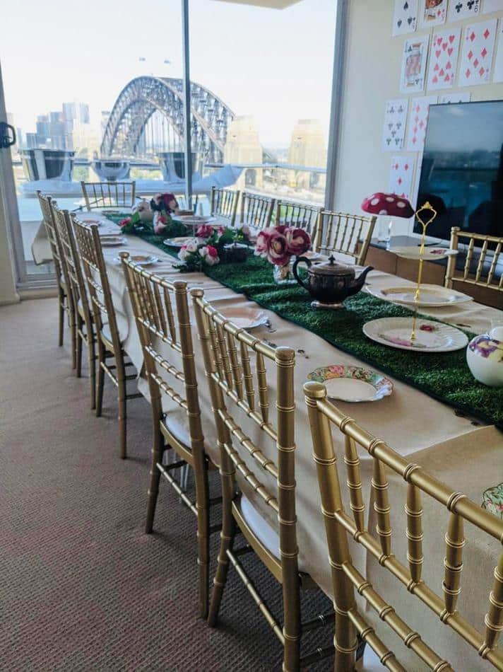 A table set up for a tea party with a view of the Sydney Harbour Bridge, featuring gold Tiffany chair hire.