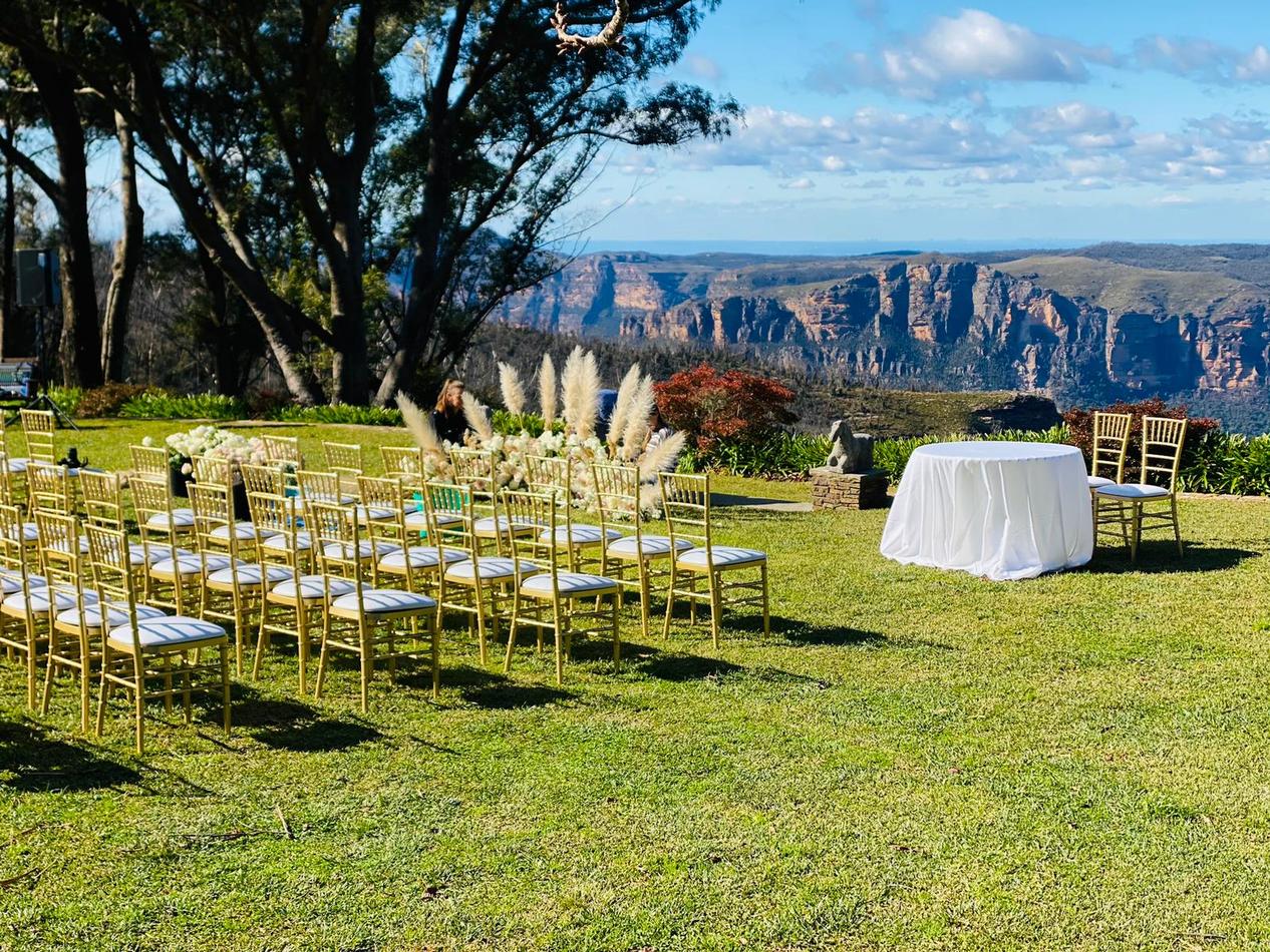 A wedding ceremony set up in a grassy area with mountains in the background, featuring Gold Tiffany Chair Hire.