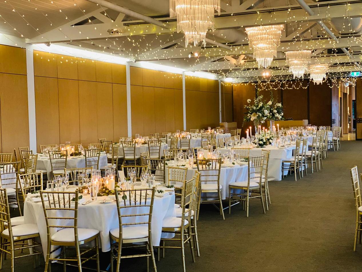 A large hall with tables and chairs set up for a wedding reception, featuring elegant gold Tiffany chair hire.