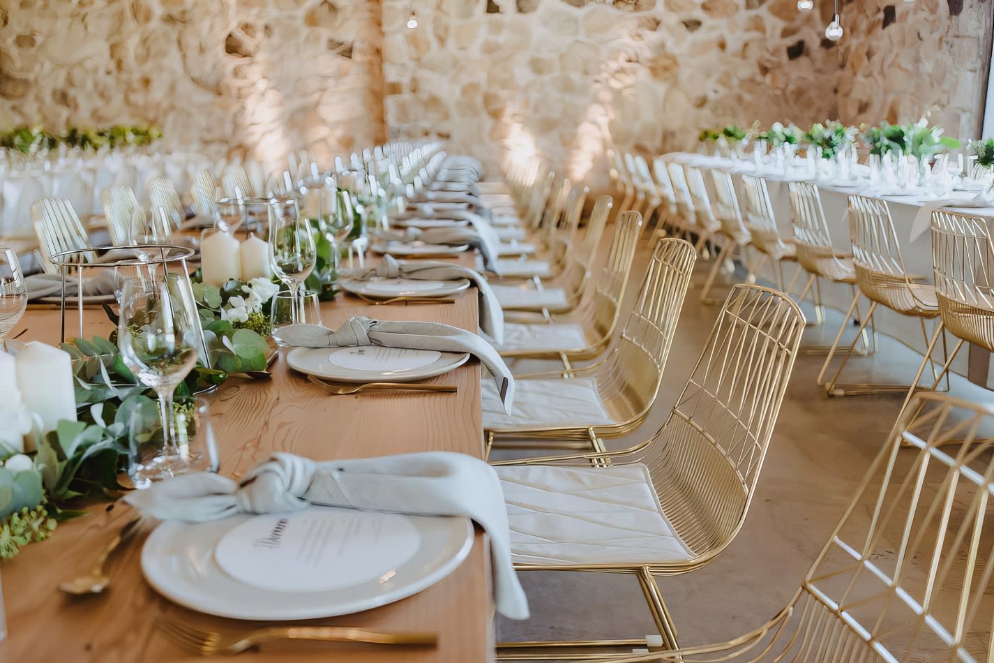 A table set up with white linens and greenery, accentuated by elegant gold wire chairs.