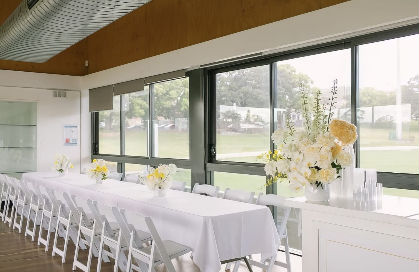 A long table is set up in a room with windows, decorated with elegant White Padded Folding Chair Hire (Gladiator chair).