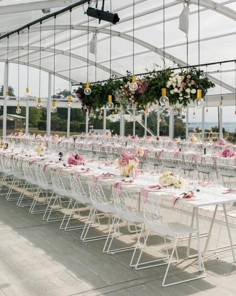 An outdoor wedding reception set up with white tables and pink flowers, featuring white wire chairs.