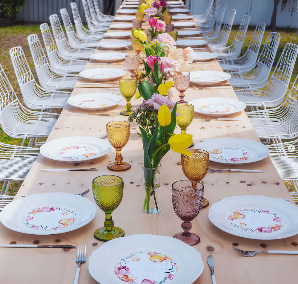 A long table set with colorful plates and glasses, featuring elegant White Wire Chairs.