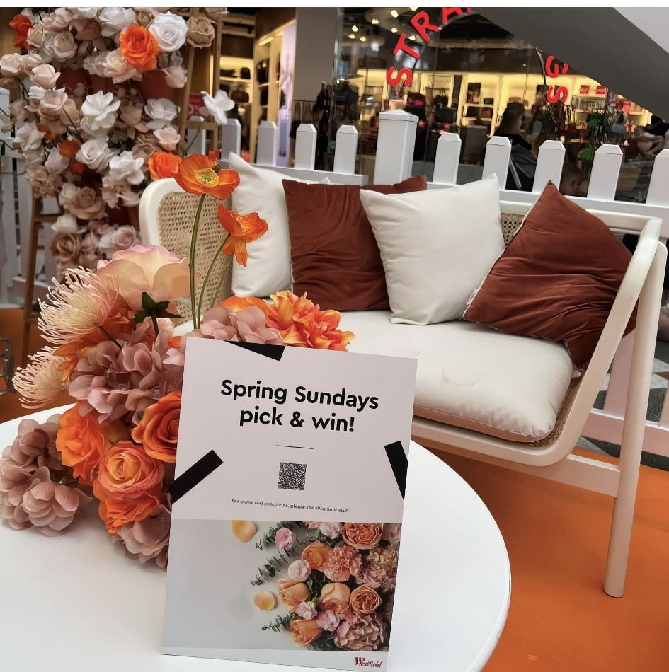 A table adorned with flowers and a sign that says "Spring Sundays," set against a backdrop of a white rattan sofa lounge.