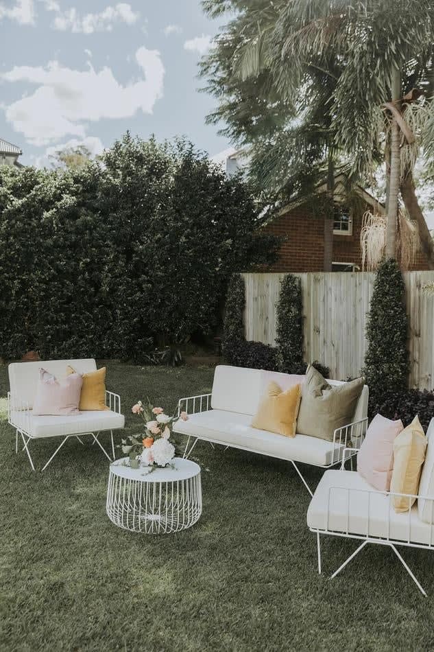 A white outdoor furniture set with wire arm chairs, hired and adorned with white velvet cushions, in a backyard.