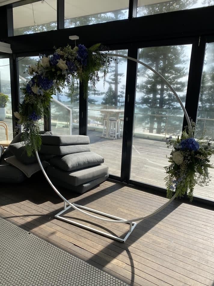 A beautiful wedding arch with flowers on it in a room with a view of the ocean, available for white hoop backdrop hire.