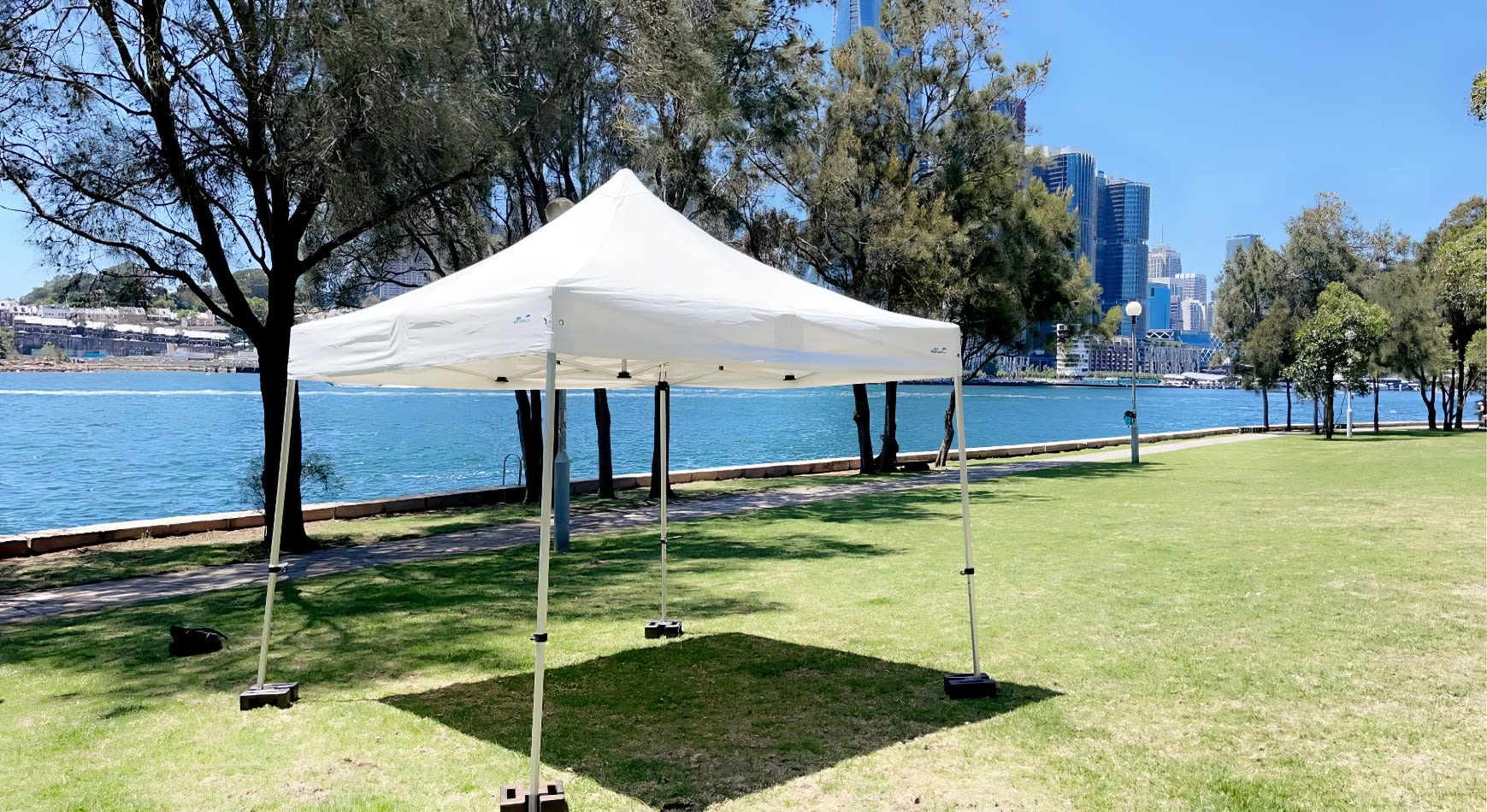 A white tent, the 3mx3m Pop Up Marquee With White Roof, is set up on a grassy area near a lake.