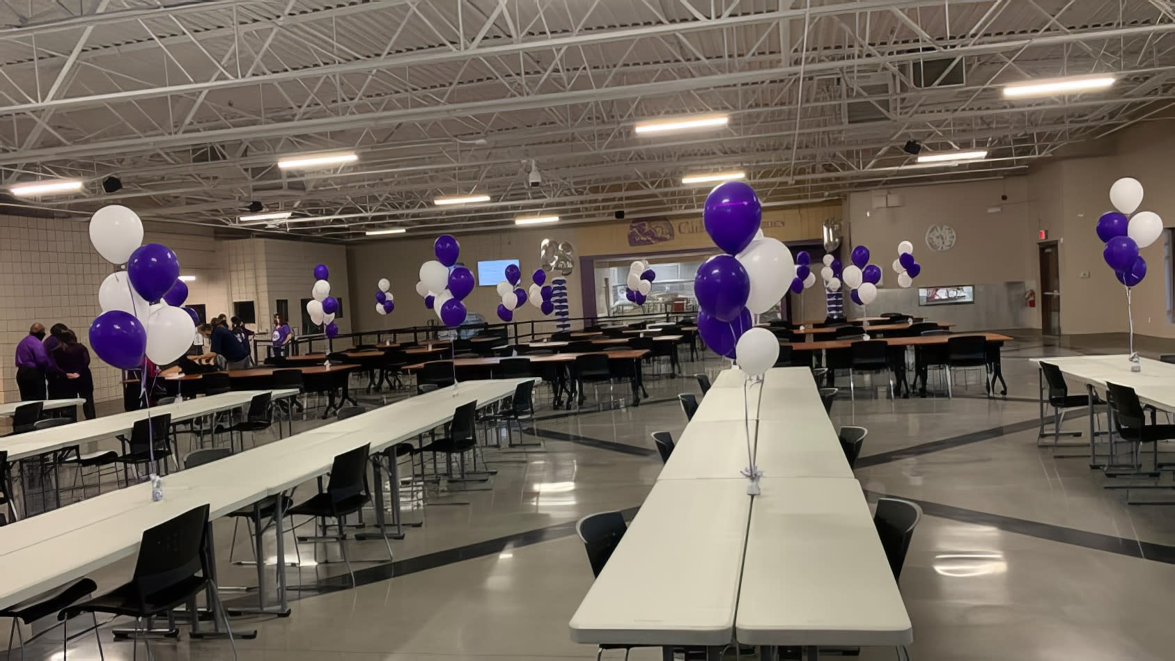 Description (modified): Purple and white balloons decorating plastic trestle tables in a school cafeteria.