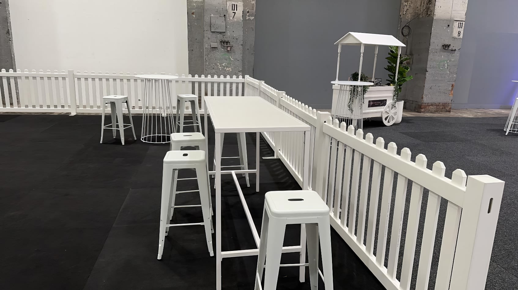 A room adorned with a white picket fence and white stools surrounding a White Rectangular Tapas Table Hire with a white top.