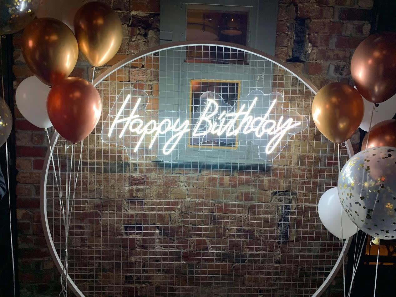 A happy birthday sign with balloons in front of a brick wall.