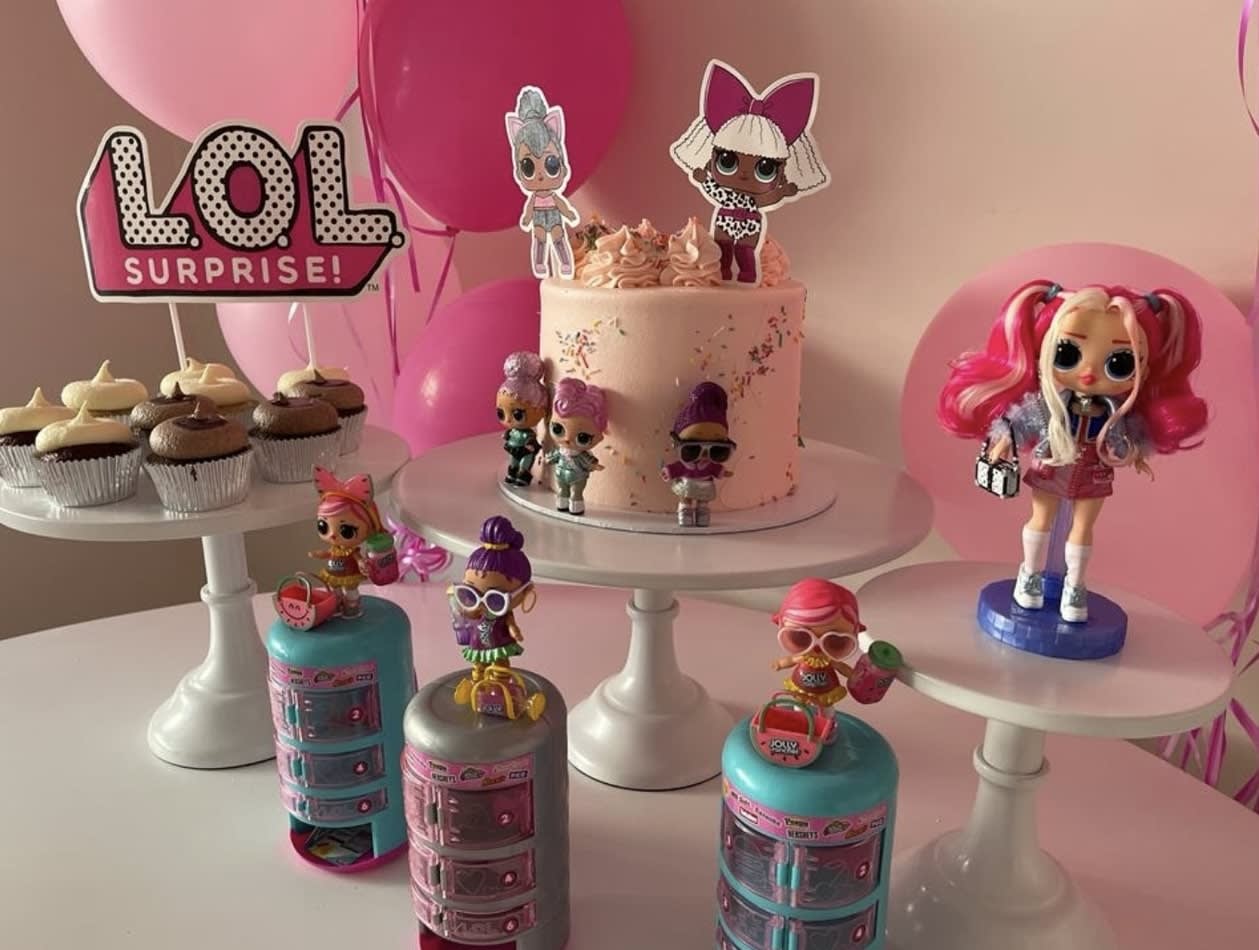 A pink cake with dolls and balloons on a white metal cake stand.