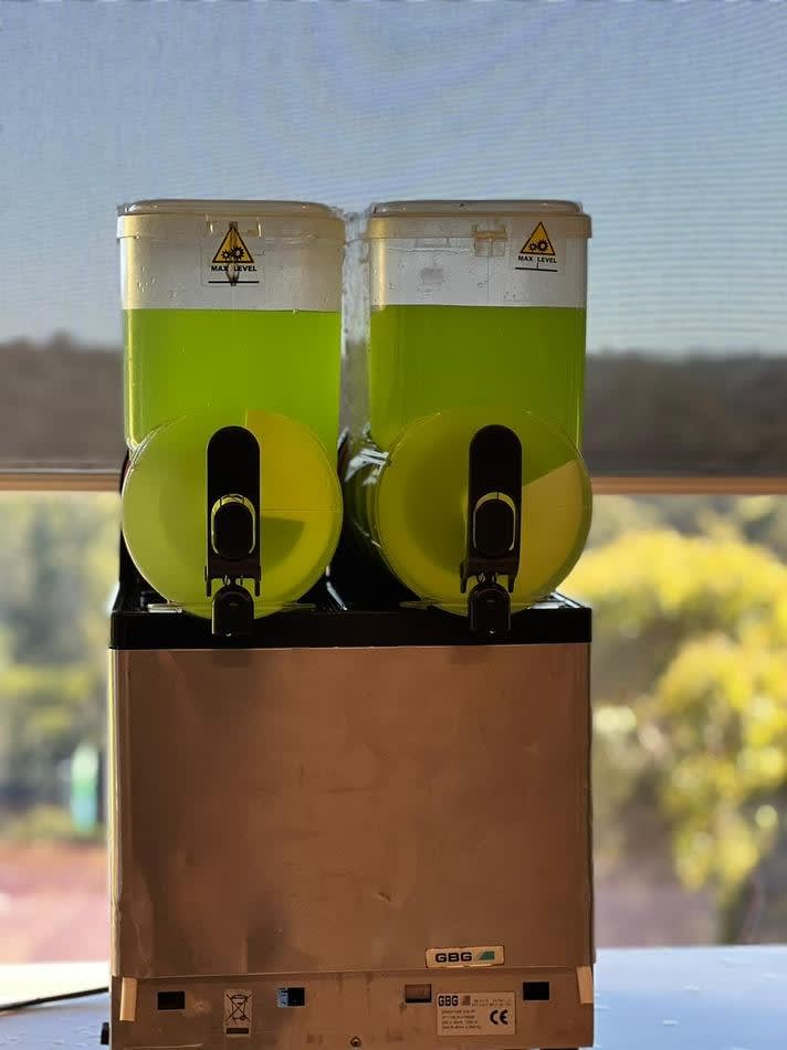 Two green juice dispensers on a table in front of a window, available for Slushie Machine Hire with a package that includes *180 Drinks*.